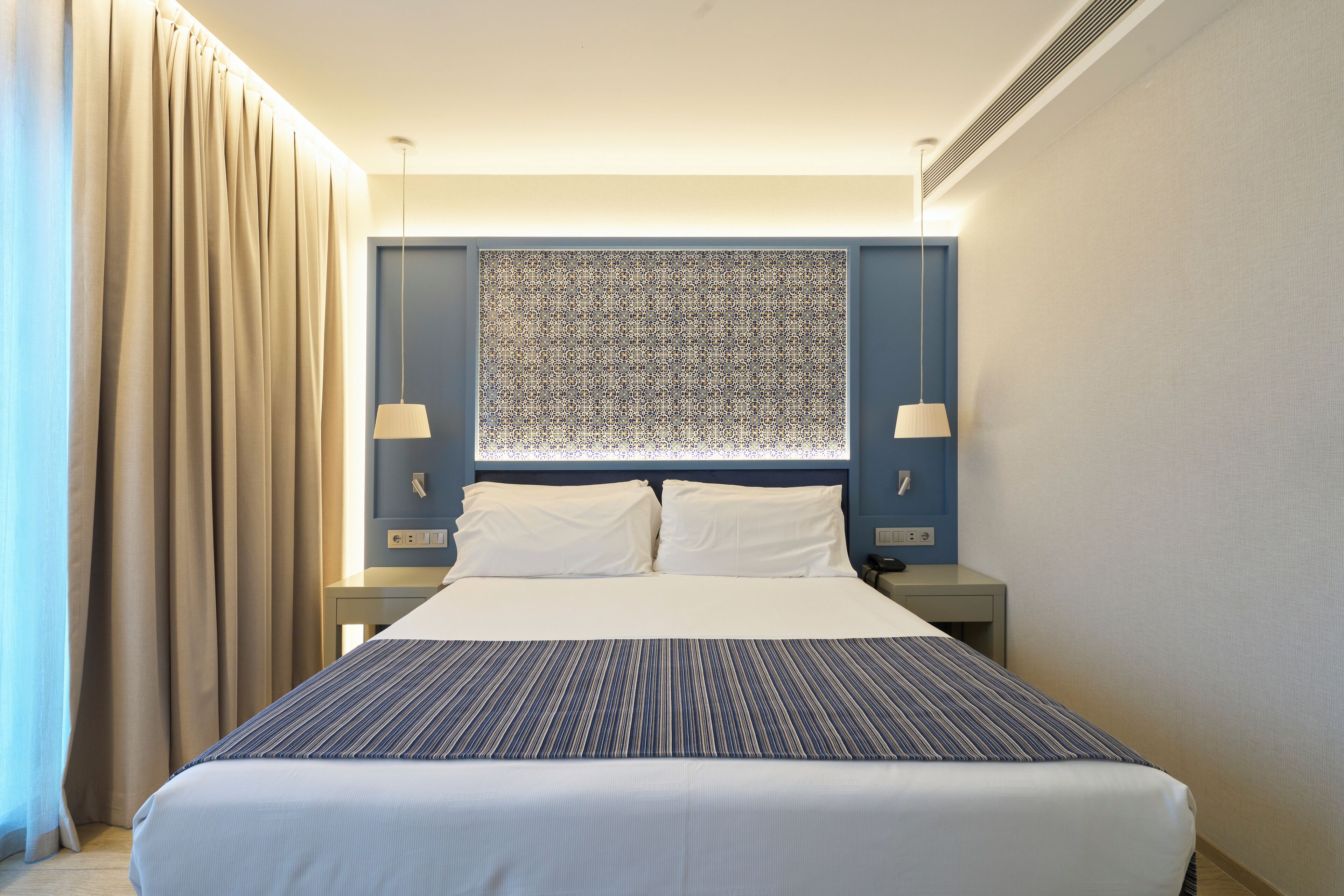 A hotel room with a large bed, two bedside tables, a nightstand, a patterned headboard, and a window with curtains.