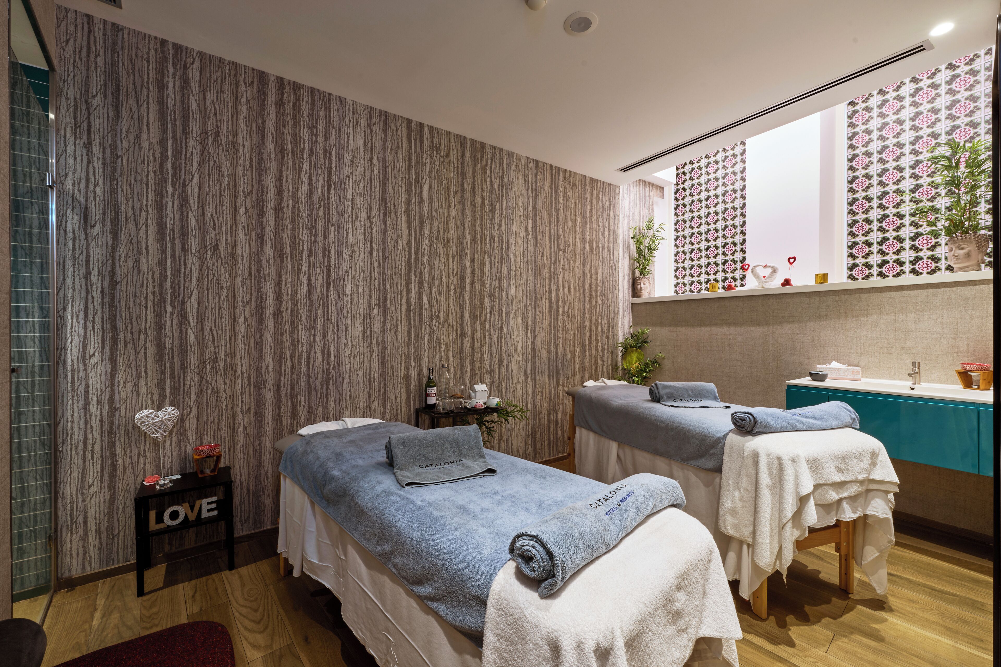 Ein Spa-Raum mit zwei Massageliegen, einem kleinen Tisch mit Flasche und herzförmiger Dekoration, sowie einer Wand mit gemustertem Wandpapier.
