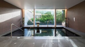A modern indoor swimming pool with clear glass walls, a metal handrail, and a view of greenery outside.