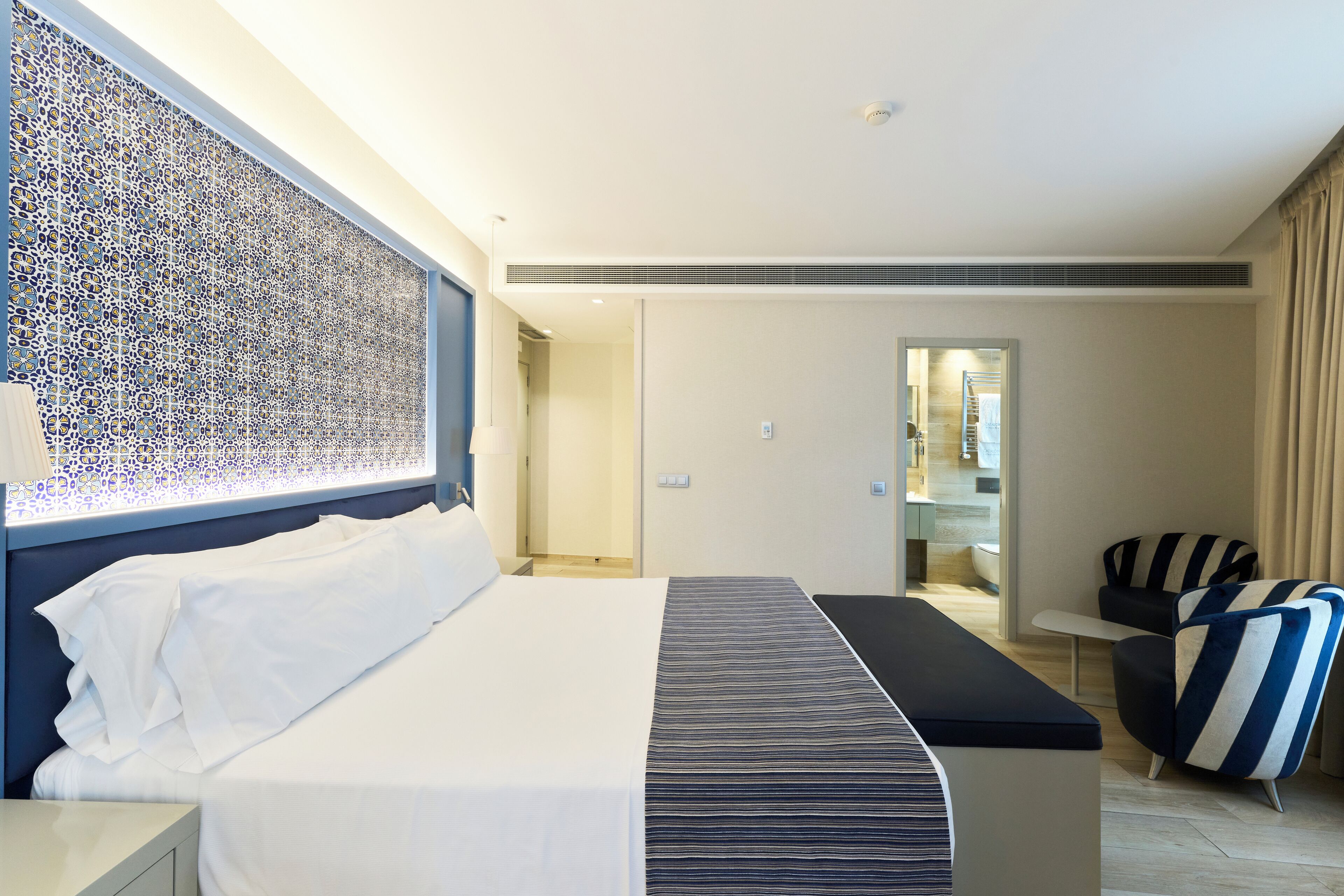 A modern hotel room with a neatly made bed, a bedside table, a chair, and a view into the bathroom.