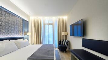 Camera d'albergo con un letto grande, una panca, una TV e vista all'esterno attraverso tende trasparenti.