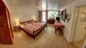 A bedroom with a wooden bed, a desk, and a chair. There is a window with curtains, a wall-mounted TV, and a shelf with plants. - Hotel Merkurwald-Restaurant Wolpertinger (Baden-Baden)