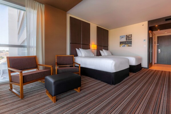 A hotel room with a large bed, two armchairs, a small ottoman, a window with curtains, and a city view. - Hilton Garden Inn Neuquen (Neuquén)