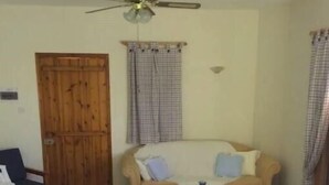 Una sala de estar con un sofá, dos sillas de madera, una mesita, un ventilador de techo y una puerta de madera.