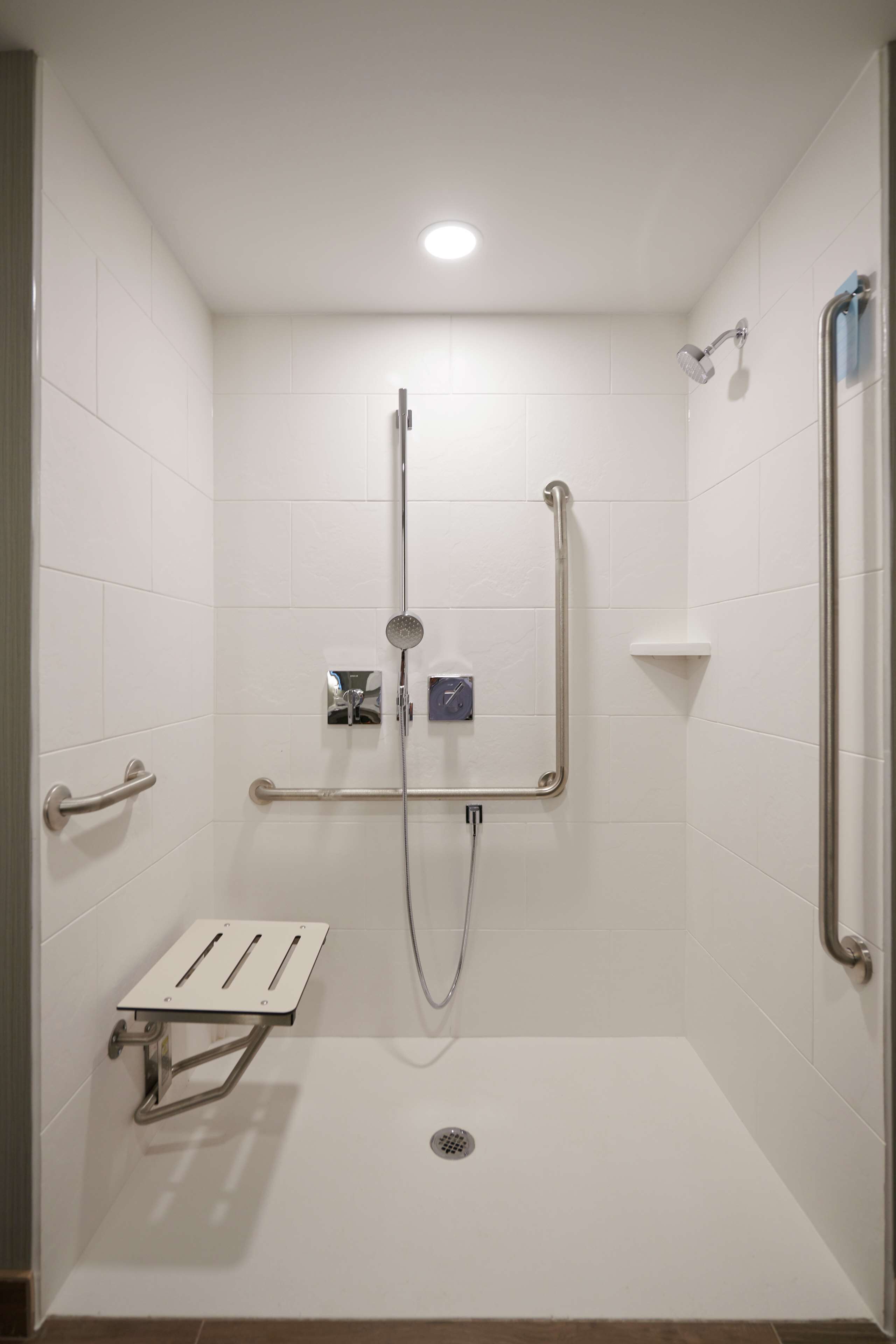 Zona de ducha con paredes de azulejos blancos, un cabezal de ducha manual, una barra de apoyo y un desagüe en el suelo.