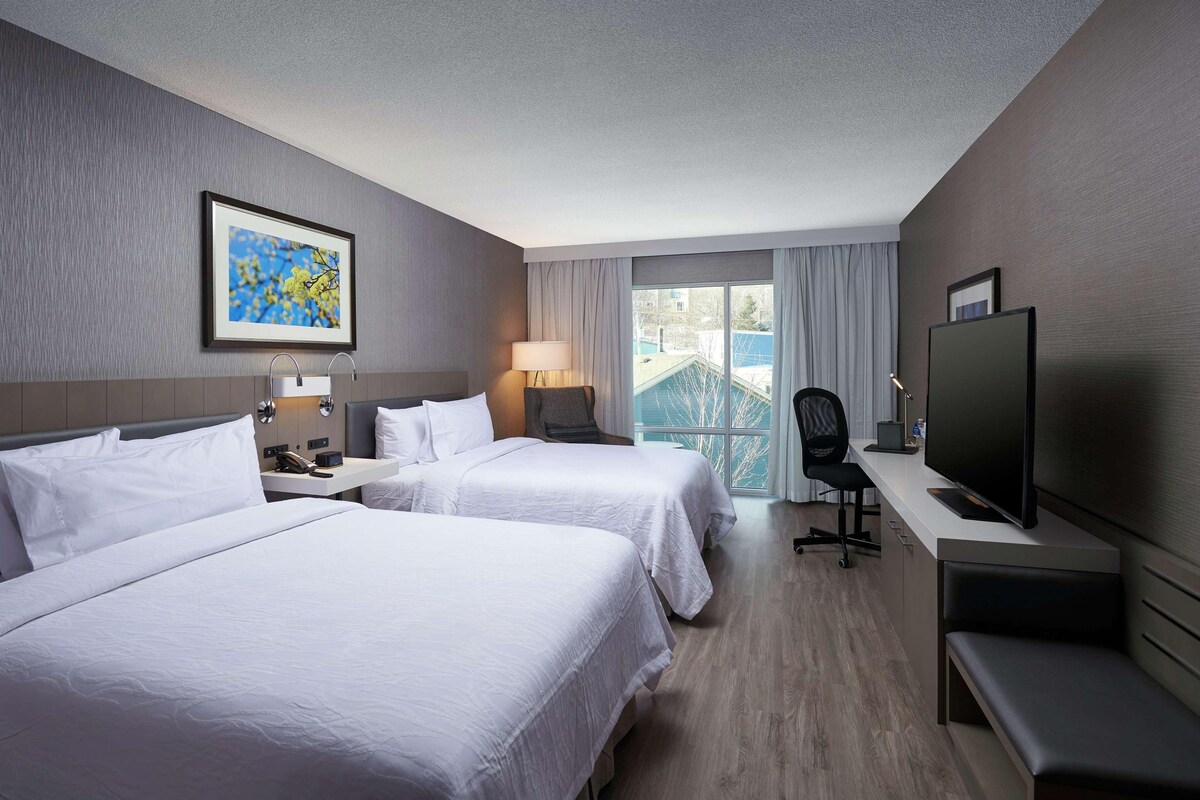 A hotel room with two beds, a desk with a computer, a TV, and a window with curtains.