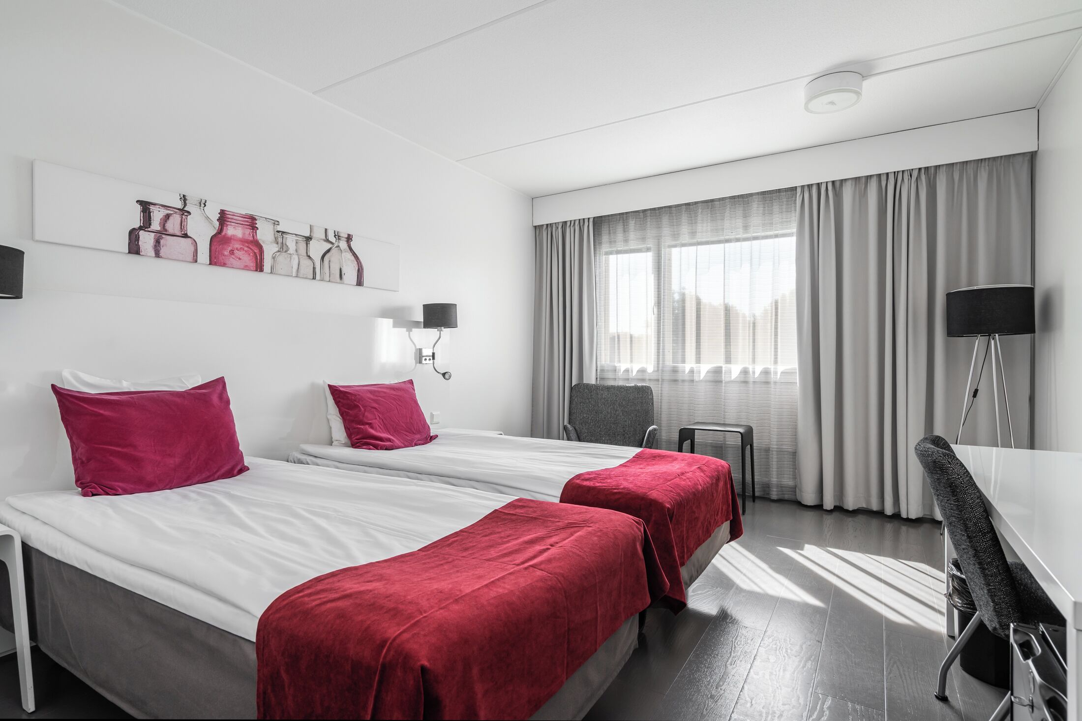 Habitación de hotel con dos camas, un escritorio, una silla, una lámpara, un ventanal con cortinas y una obra de arte en la pared.