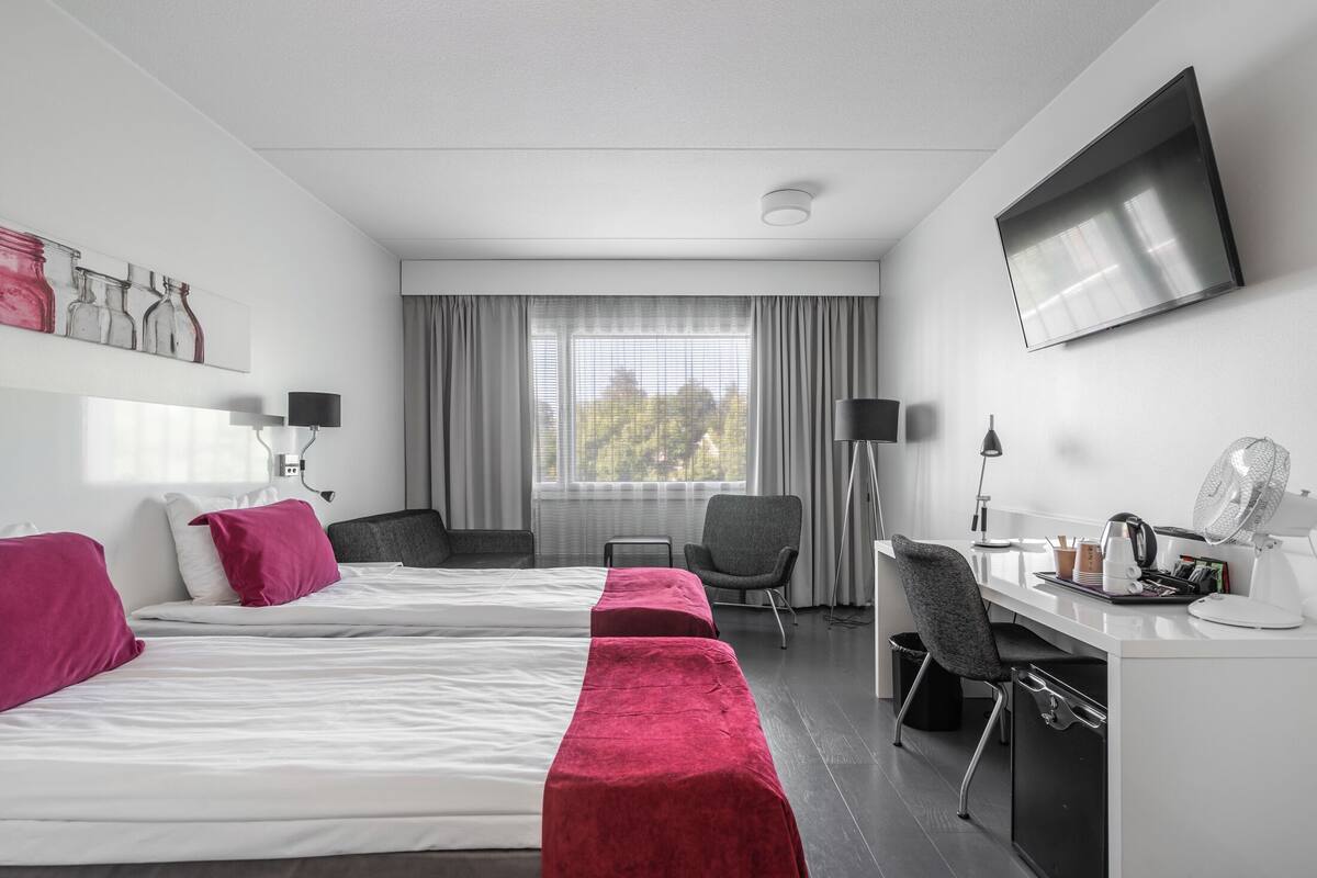 Ein Hotelzimmer mit zwei Betten, einem Schreibtisch, einem Stuhl, einem Fernseher und einem Fenster mit Vorhängen.