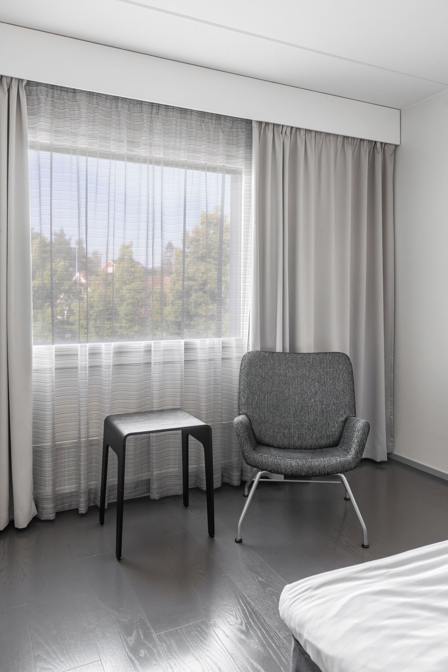 A modern hotel room with a large window, a grey upholstered chair, a small table, and a bed.