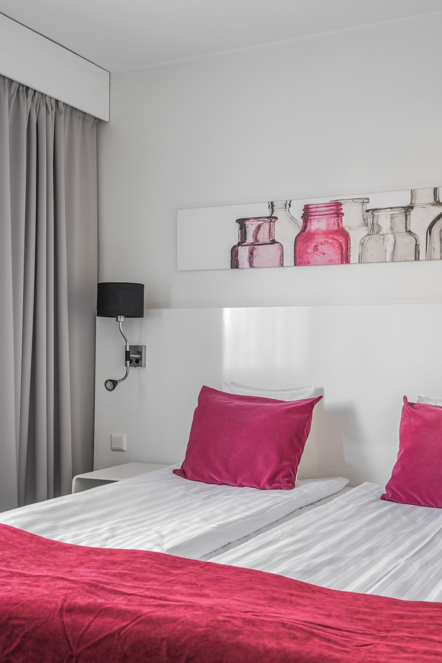 Ein Hotelzimmer mit Bett, Kissen, einer Wandlampe und einem Regal mit Glasflaschen.