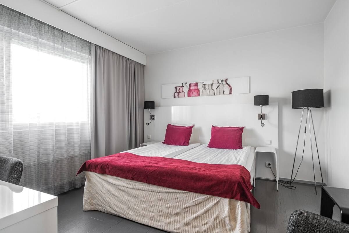 Ein modernes Hotelzimmer mit Bett, Nachttischen, einem Schreibtisch, einem Stuhl, einer Lampe und einem Vorhangfenster.