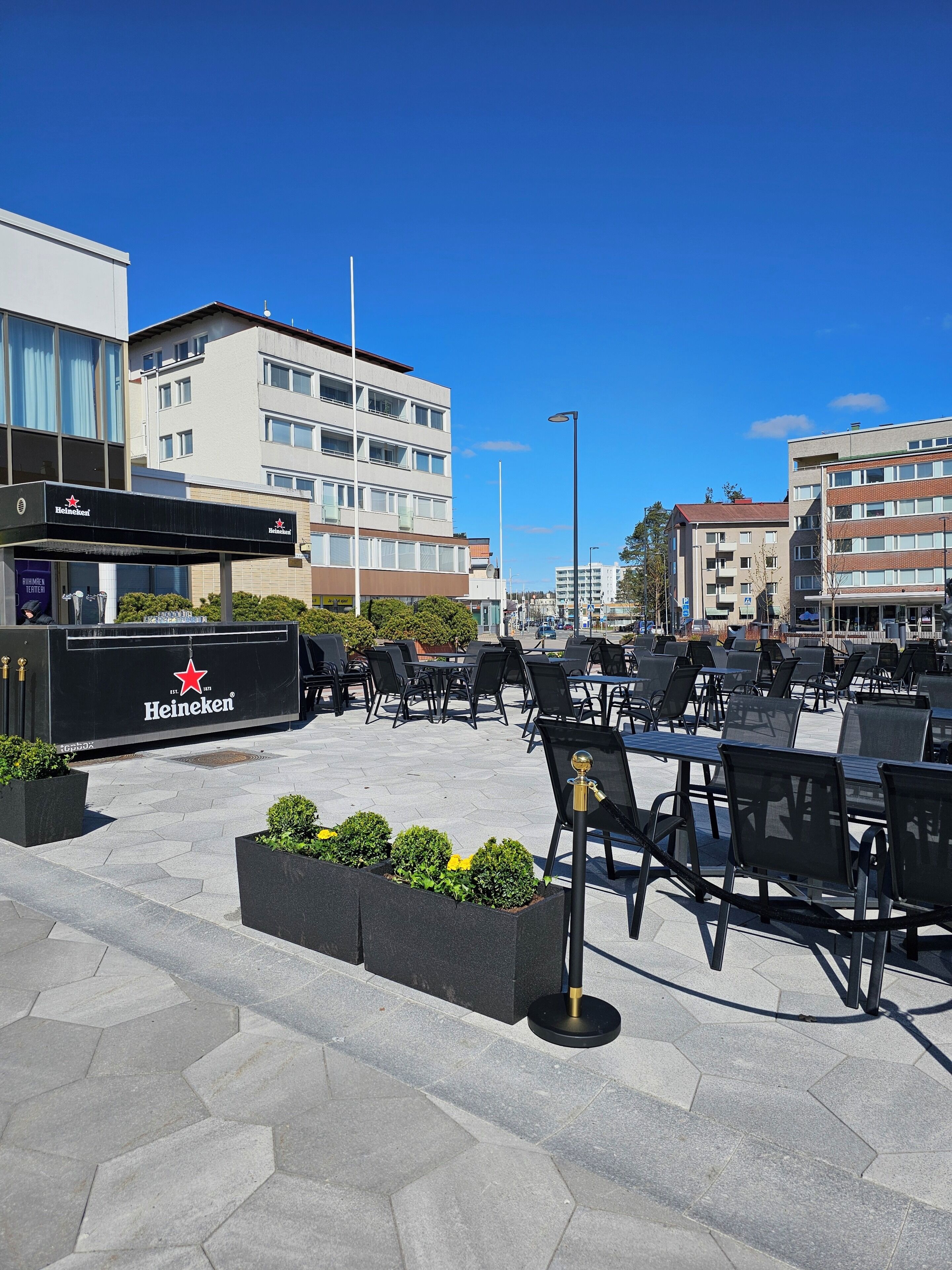 An outdoor seating area with hexagonal stone tiles, black metal chairs, and a Heineken branded kiosk.