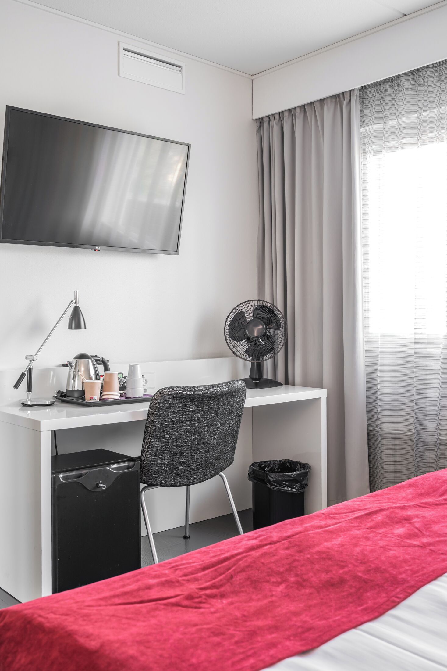 Una habitación de hotel con una cama, un escritorio con una silla, un televisor, un ventilador y una ventana con cortinas.