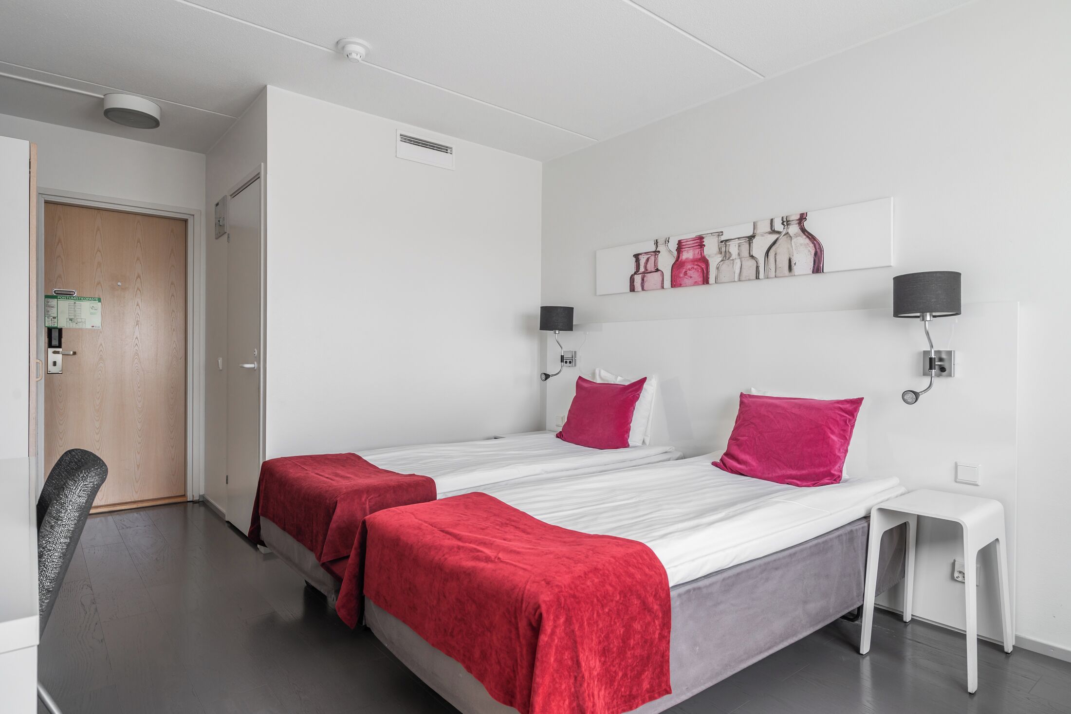 Quarto de hotel com duas camas, uma escrivaninha, uma cadeira, uma mesinha e uma luminária na parede.