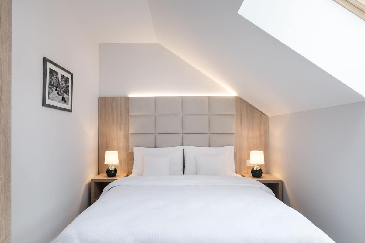A modern bedroom with a sloped ceiling, a large bed with white linens, two bedside tables with lamps, and a framed picture on the wall.