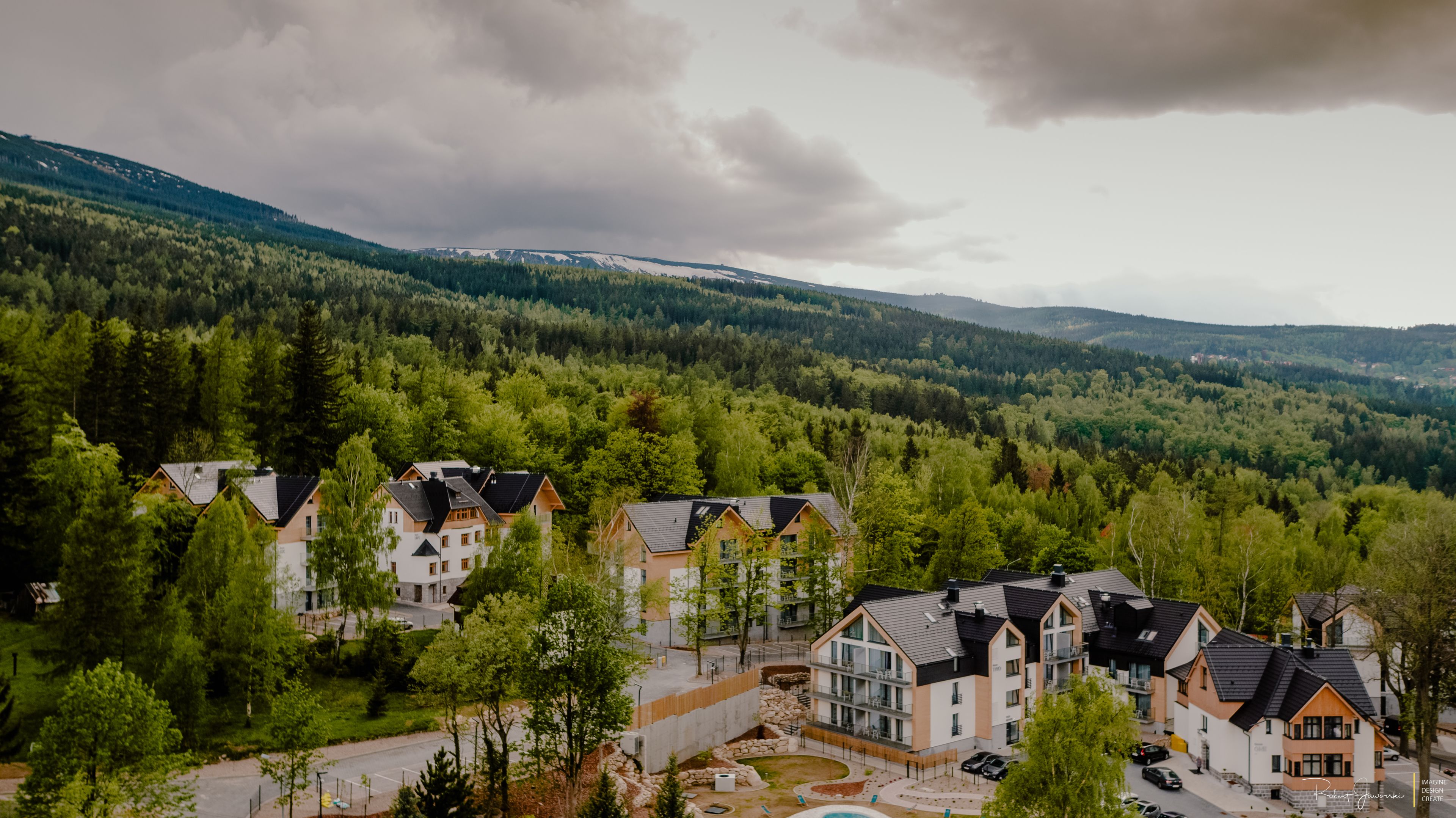 A residential area with multiple two-story houses, surrounded by dense forest and mountains in the background.