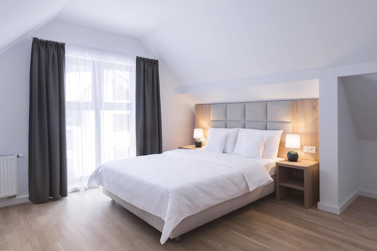A modern hotel room with a large bed, two bedside tables with lamps, a radiator, and a view of the outdoors.