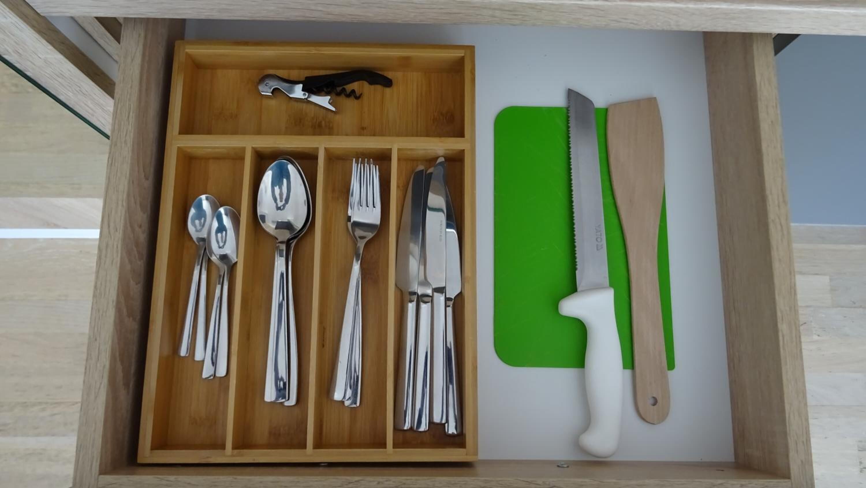 A drawer containing various utensils including spoons, forks, and knives, along with a green cutting board and a knife with a white handle.