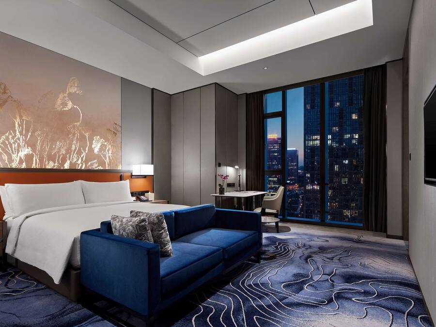 Ein modernes Hotelzimmer mit einem großen Bett, einem blauen Sofa, Blick auf die Stadt und einem großen Kunstwerk an der Wand.