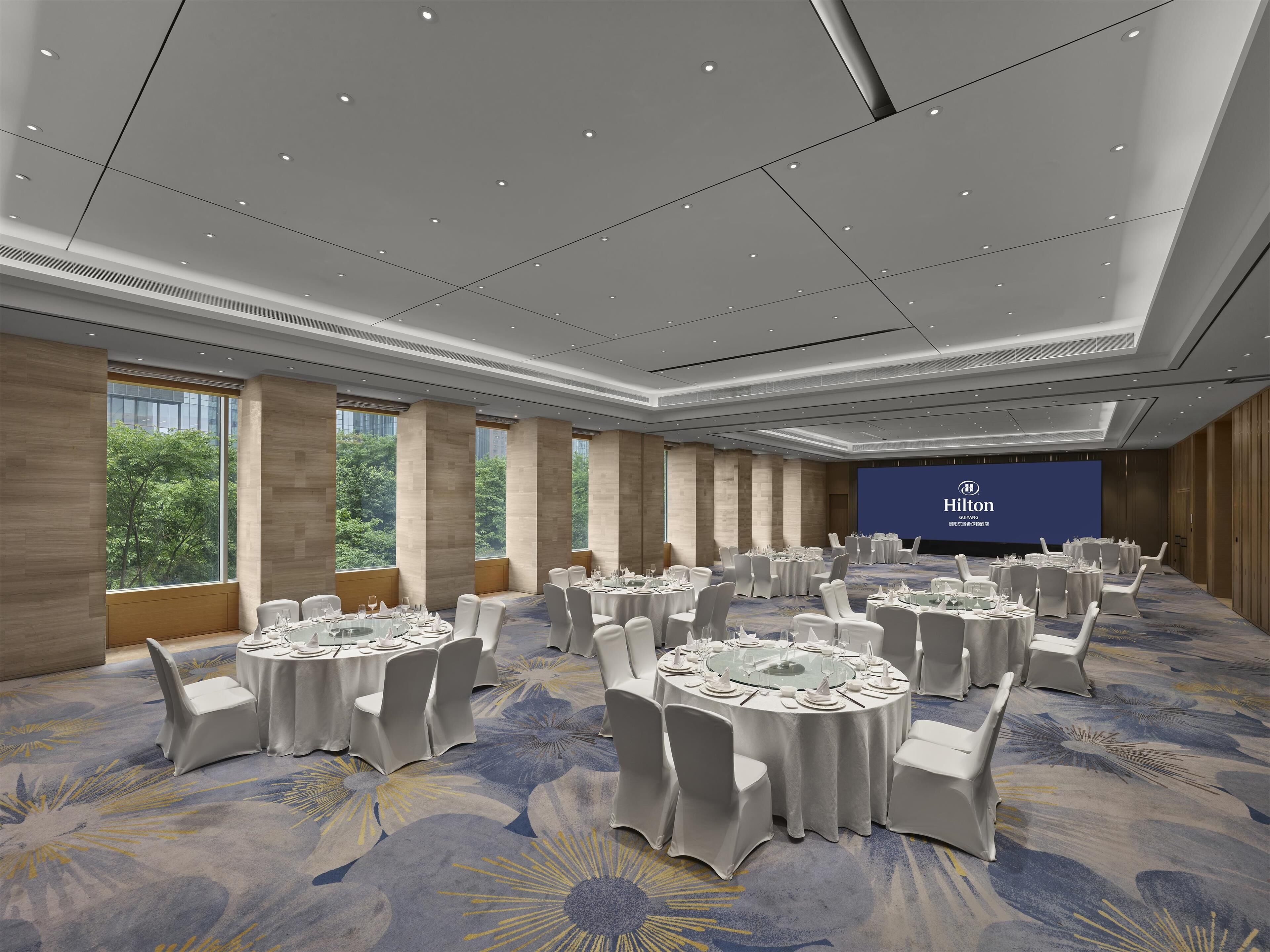 A conference room with round tables, white chairs, and a large screen displaying the Hilton logo.
