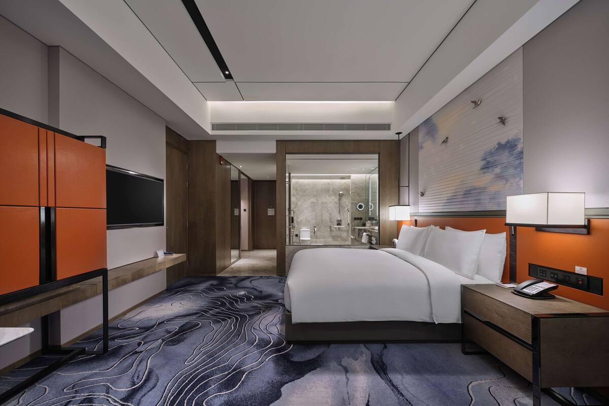 Une chambre d’hôtel moderne avec un grand lit, un bureau avec un téléphone, une lampe de chevet et un téléviseur sur le mur de gauche.