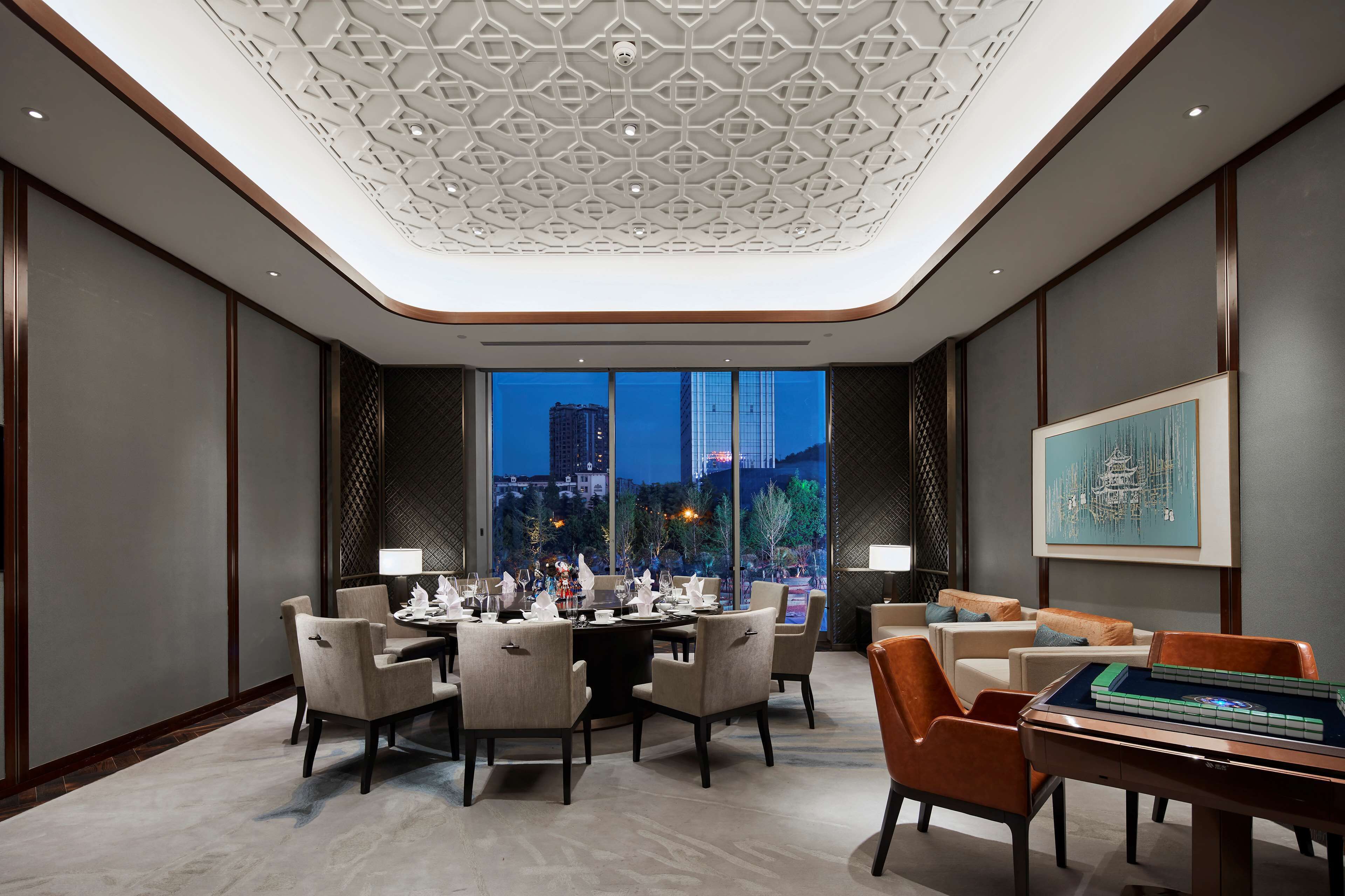 A modern dining area with a high ceiling, large windows, and a view of the cityscape at dusk.