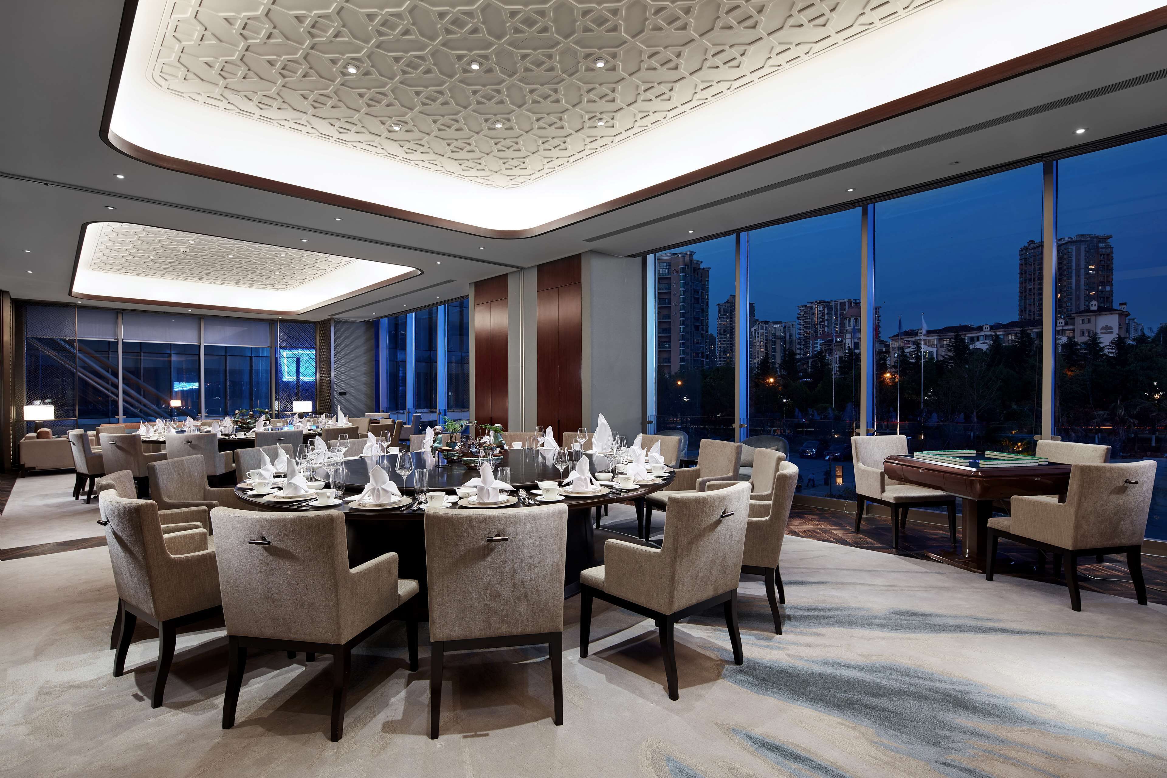 Un comedor moderno con mesas redondas dispuestas para una comida, amplias ventanas que ofrecen vistas a la ciudad y un techo decorativo.