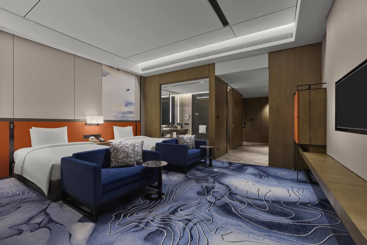 Une chambre d’hôtel avec un grand lit, deux fauteuils bleus, une petite table, un téléviseur et une salle de bain avec un miroir et une douche.