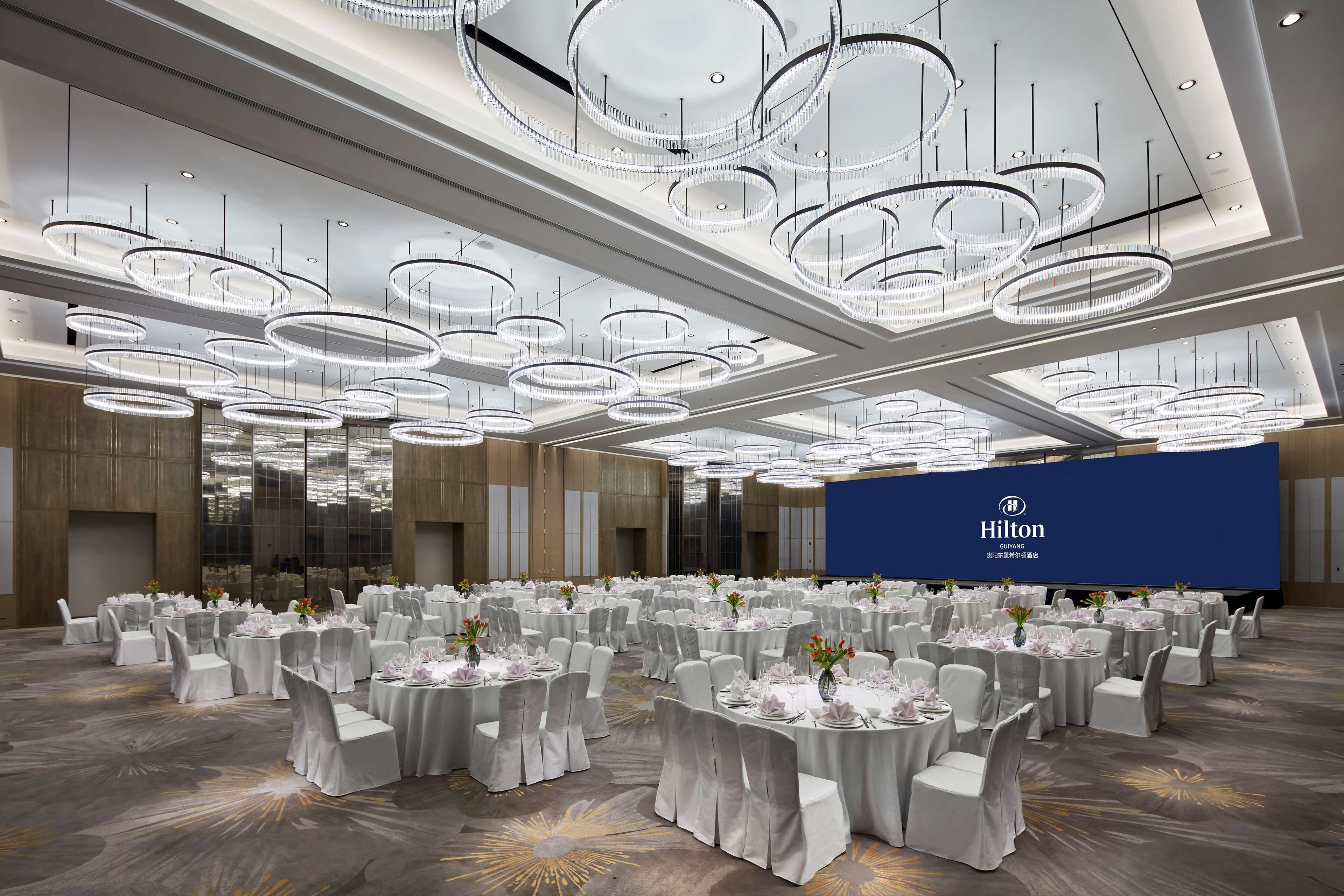 A spacious conference room with multiple round tables, white chairs, and a large blue screen displaying the Hilton logo.
