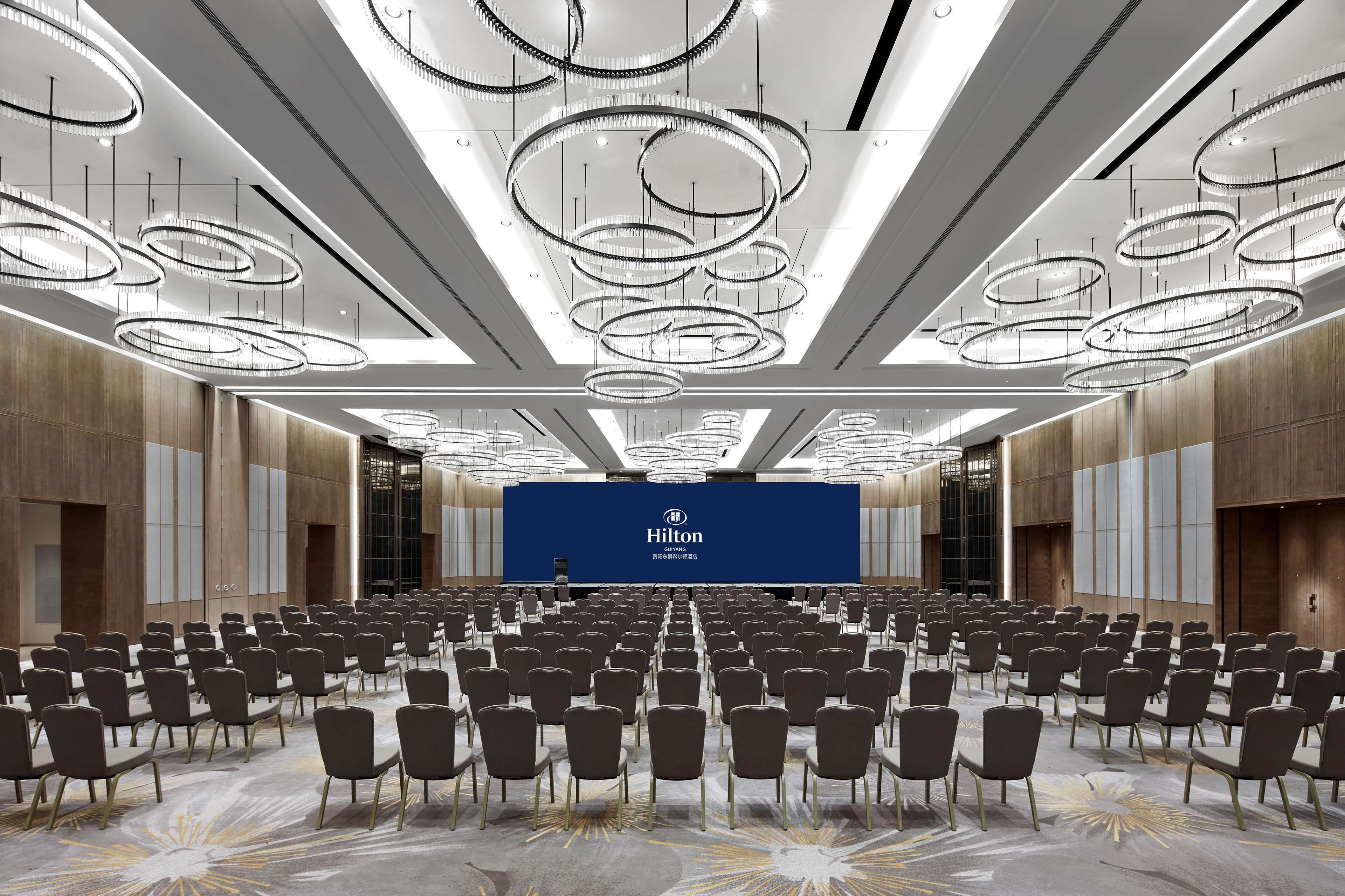 A large conference room with rows of chairs, a blue stage with Hilton branding, and modern circular chandeliers.