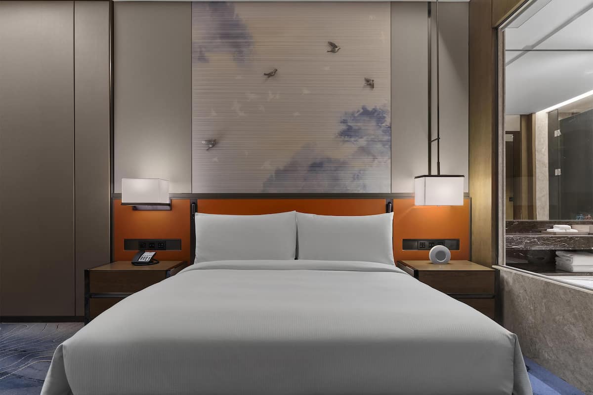Une chambre d’hôtel avec un grand lit, deux tables de chevet avec des lampes, une œuvre d’art accrochée au mur et un grand miroir.