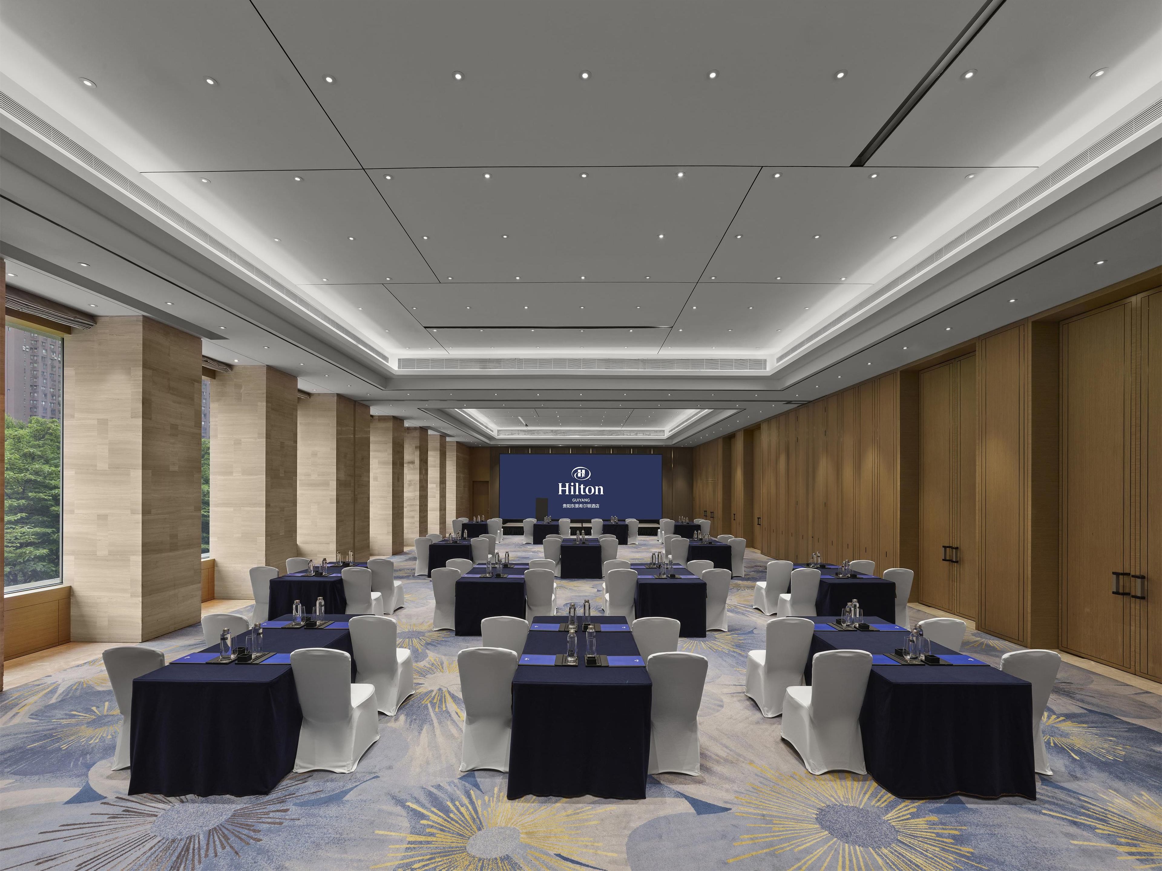 Ein Konferenzraum mit einer großen Leinwand, auf der die Marke "Hilton" zu sehen ist, mit Tischen und Stühlen in Reihen und einem Teppichboden.