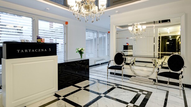 A lobby with a reception desk labeled "TARTACZNA 3", a chandelier, a large mirror, and a checkered floor.