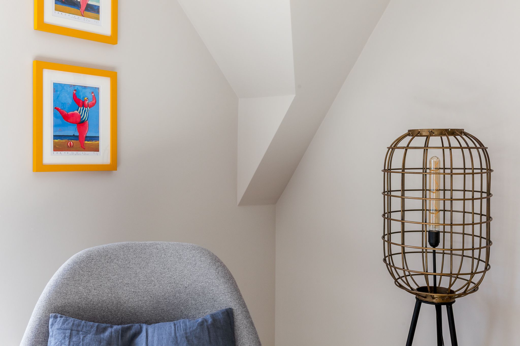 Una habitación con un sillón gris, una lámpara de pie con estructura metálica y dos obras de arte enmarcadas en la pared.