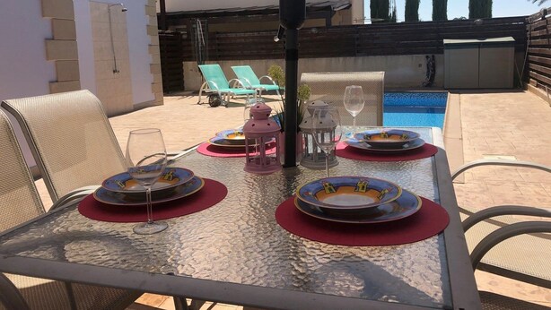 Ein Glastisch ist für das Essen gedeckt, mit Tellern, Gläsern und einer Laterne, mit Blick auf einen Swimmingpool und ein Gebäude mit einem Ziegeldach.