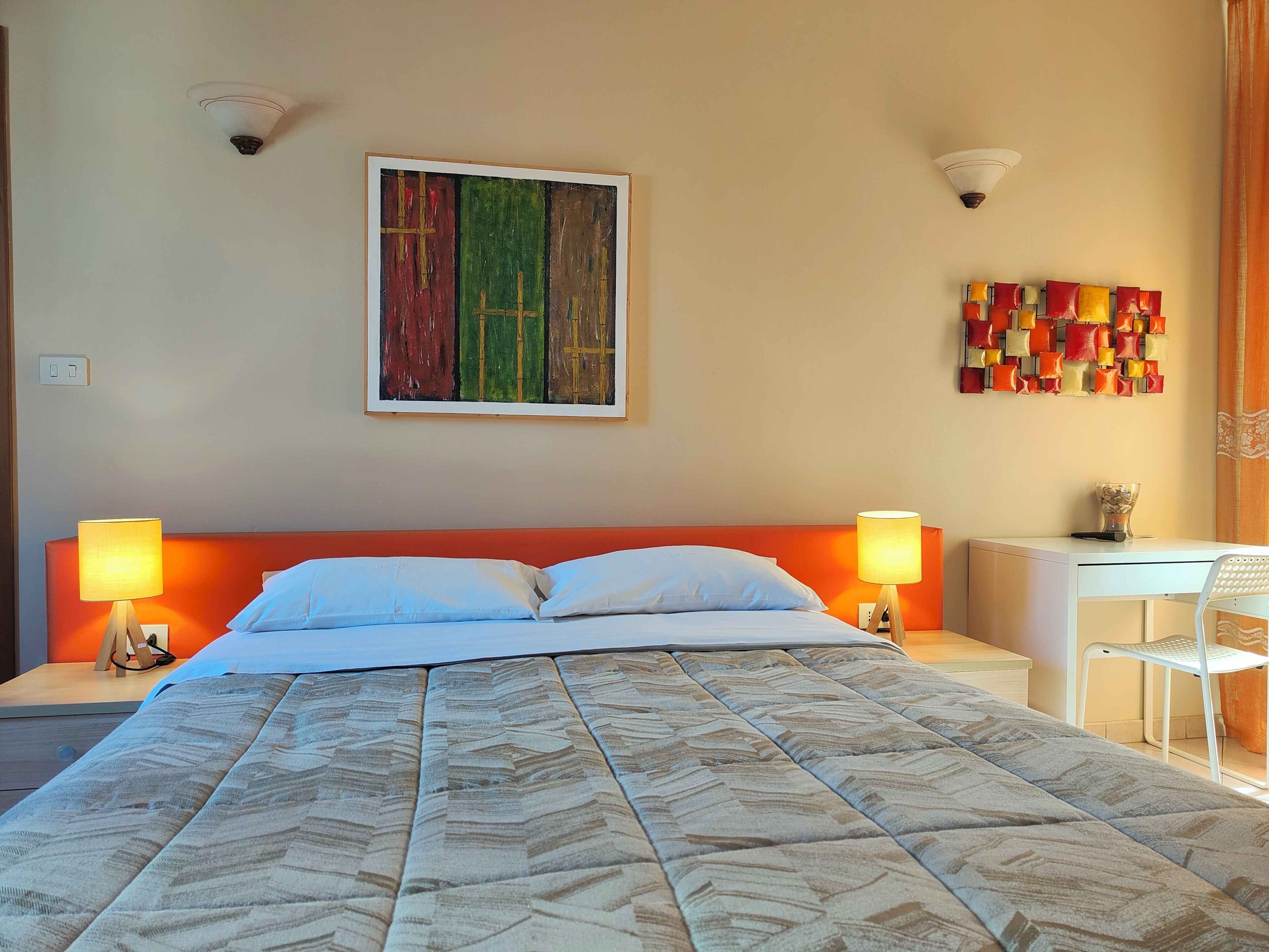 A hotel room with a bed, two bedside lamps, a desk, a chair, a wall art piece, and a wall-mounted light fixture.