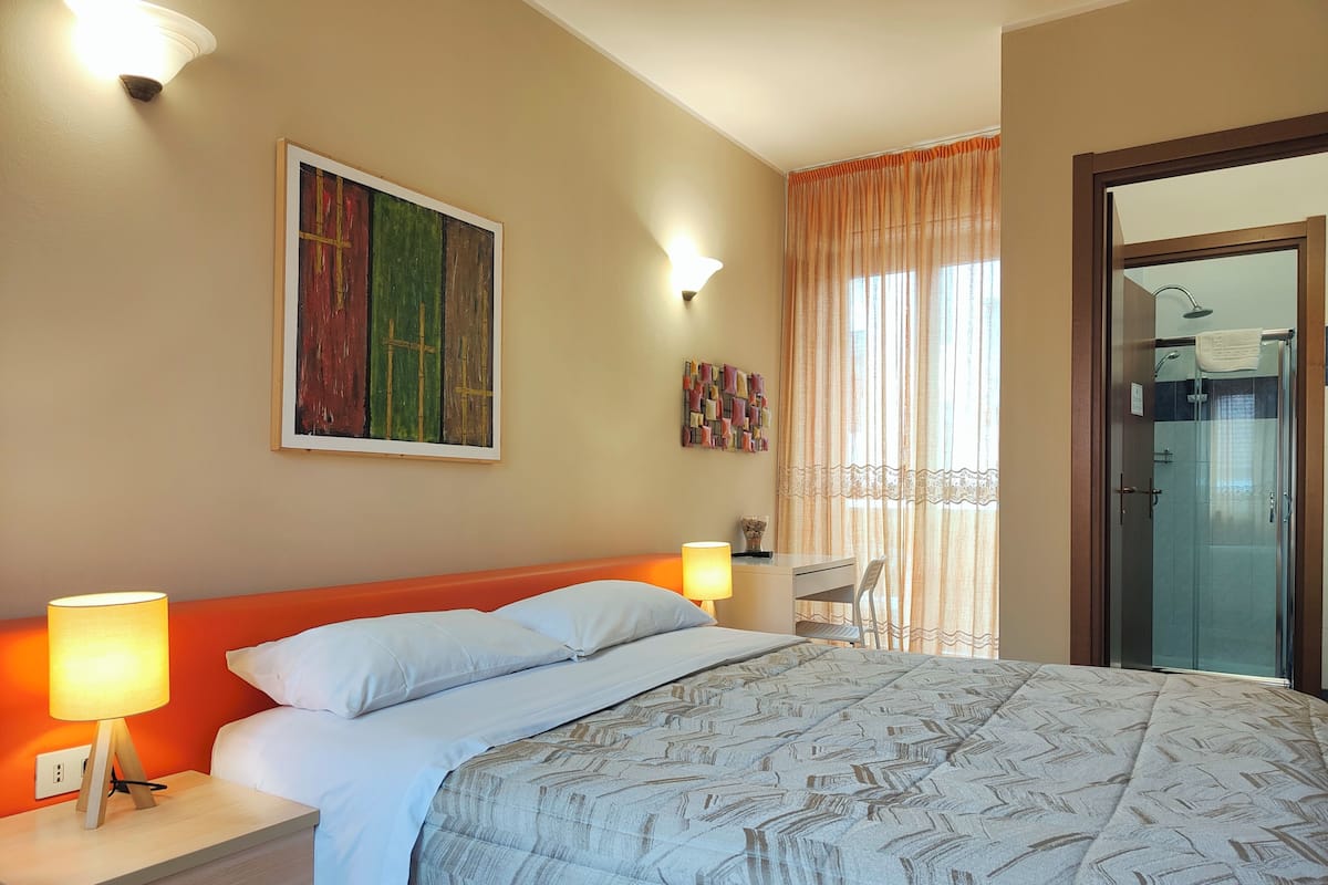 A hotel room with a bed, bedside lamps, a desk, a chair, a shower, and a painting on the wall.
