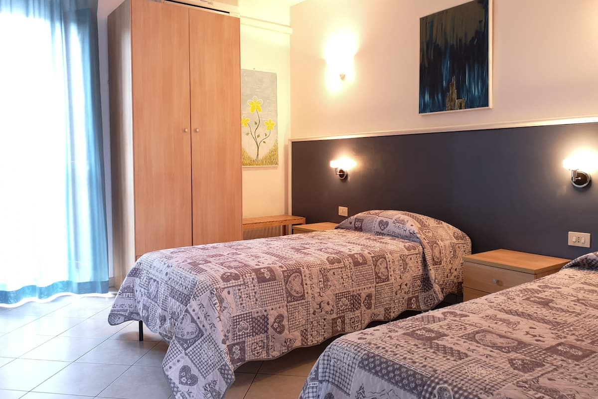 A bedroom with two beds, a wardrobe, a painting, and a wall-mounted light fixture.