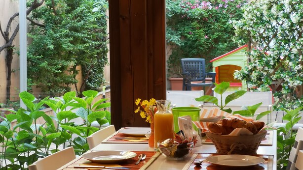 Une table dressée pour deux personnes, avec des assiettes, des couverts, un verre de jus d’orange, un panier de pain et un vase rempli de fleurs.