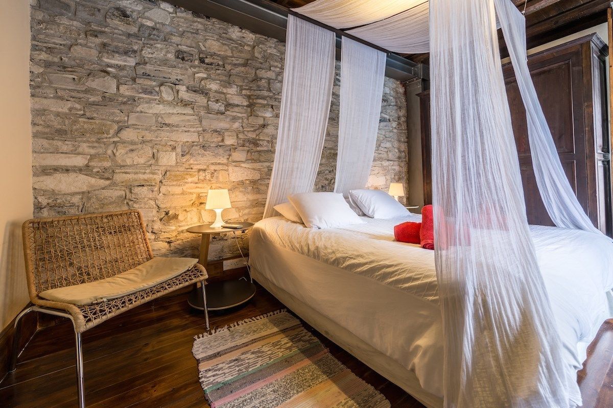 Un dormitorio con pared de piedra, una cama con ropa de cama blanca y almohadas rojas, una silla de mimbre y una pequeña mesita de noche con lámpara.