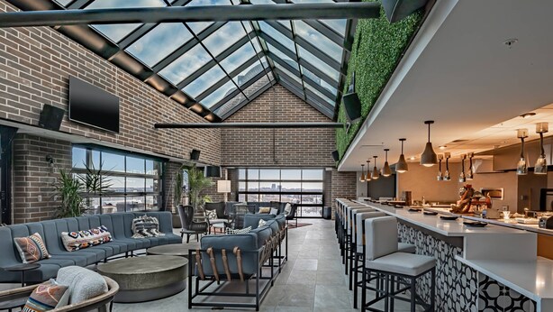 A spacious indoor area with a high ceiling, a glass roof, and a brick wall. There is a seating area with sofas and chairs, a bar area with stools, and a kitchen visible in the background. - SpringHill Suites by Marriott Fort Worth Historic Stockyards (Fort Worth)