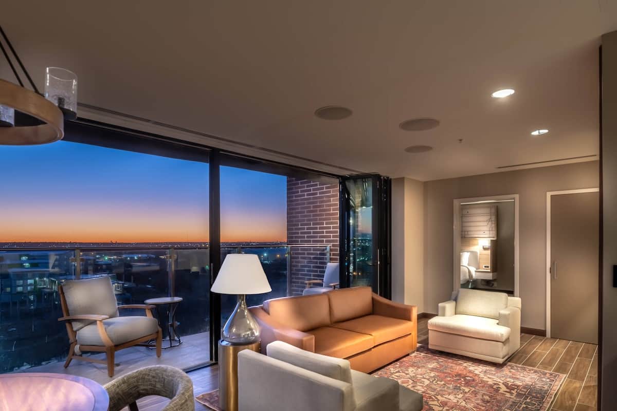 Une chambre d’hôtel moderne dotée d’un grand balcon, d’un canapé, d’un fauteuil, d’une petite table et d’une lampe.