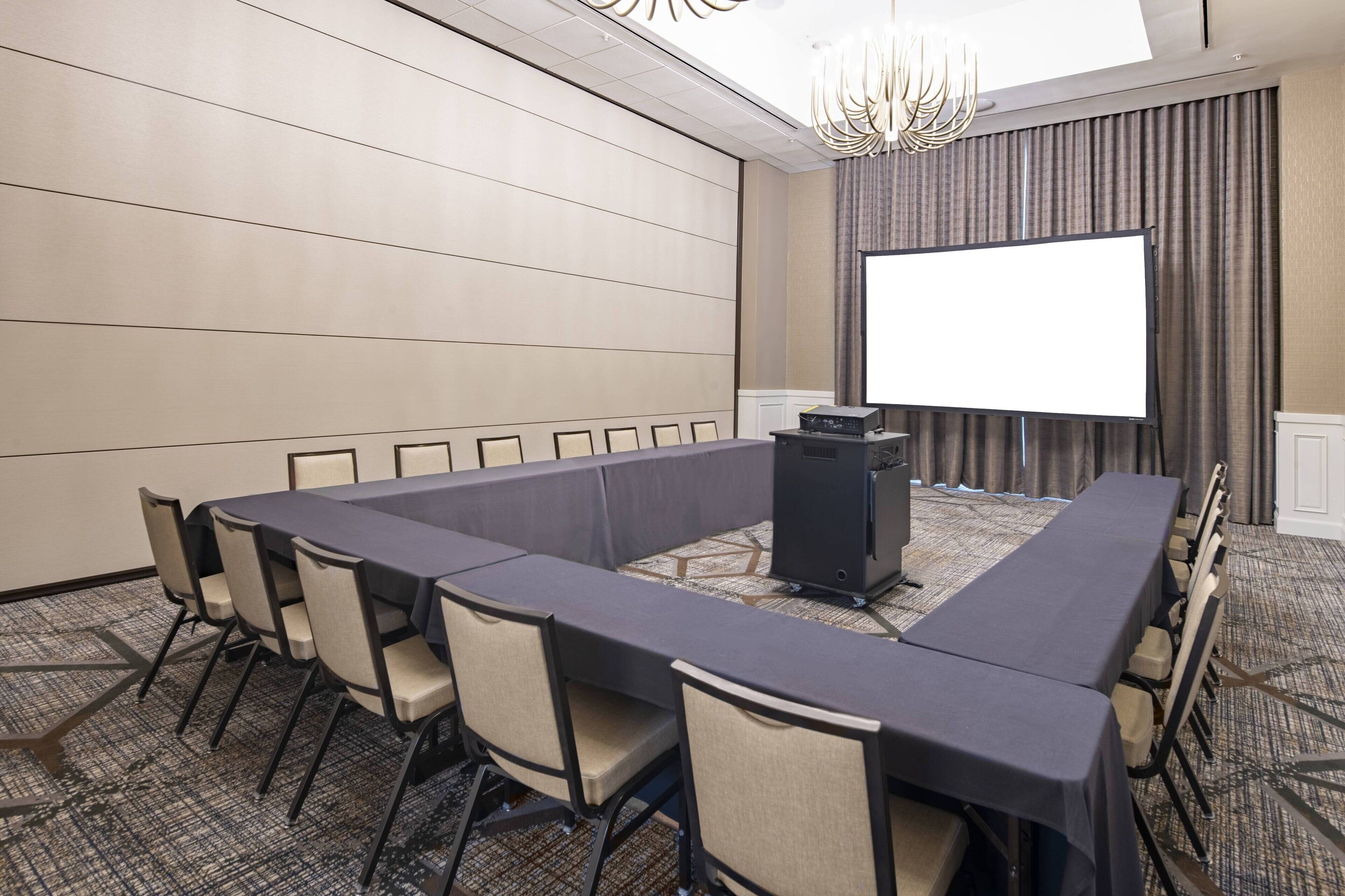 Una sala de conferencias con una larga mesa, sillas, una pantalla de proyector y piso alfombrado.