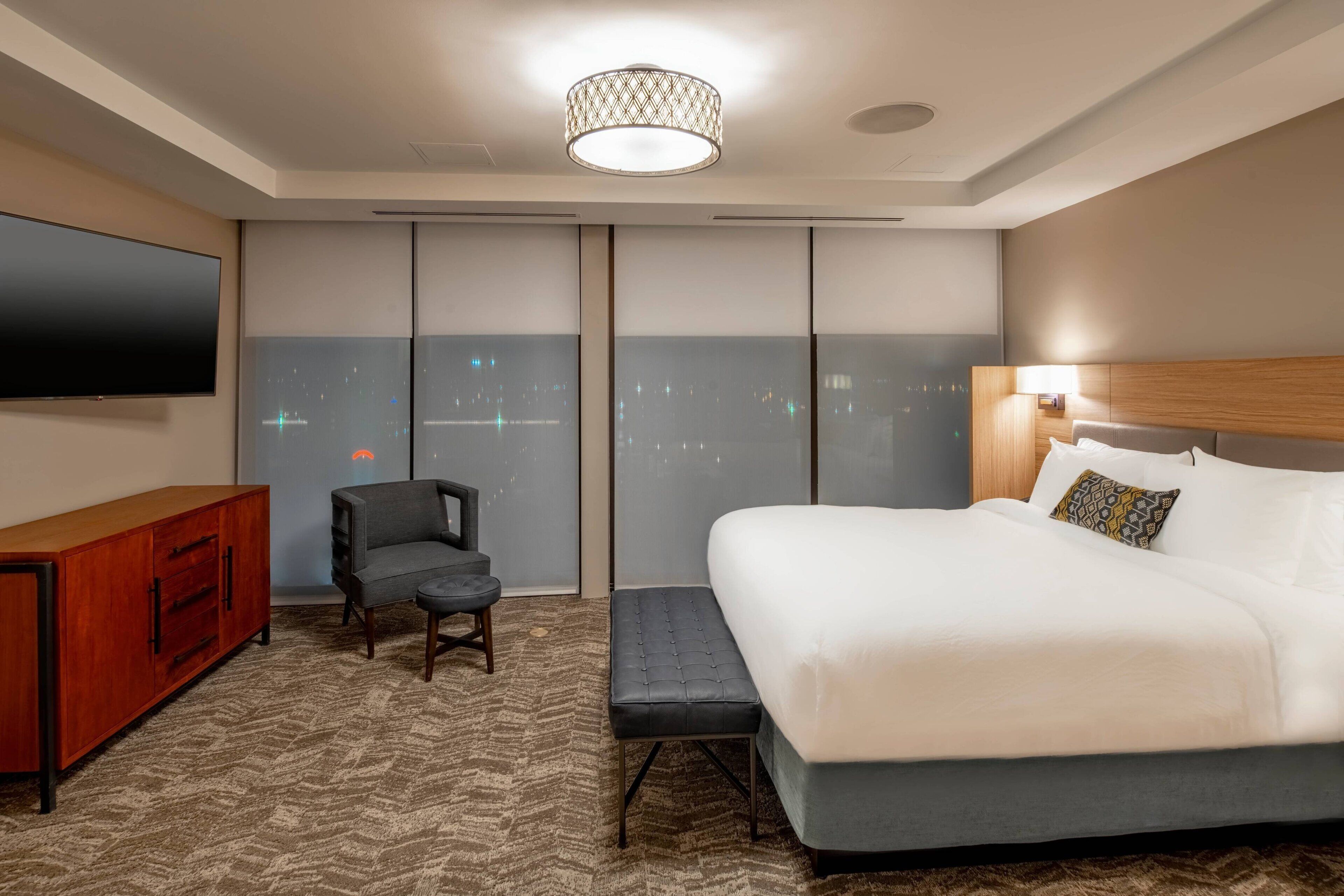 A hotel room with a large bed, a television, a seating area with a chair and ottoman, and a city view through the windows.