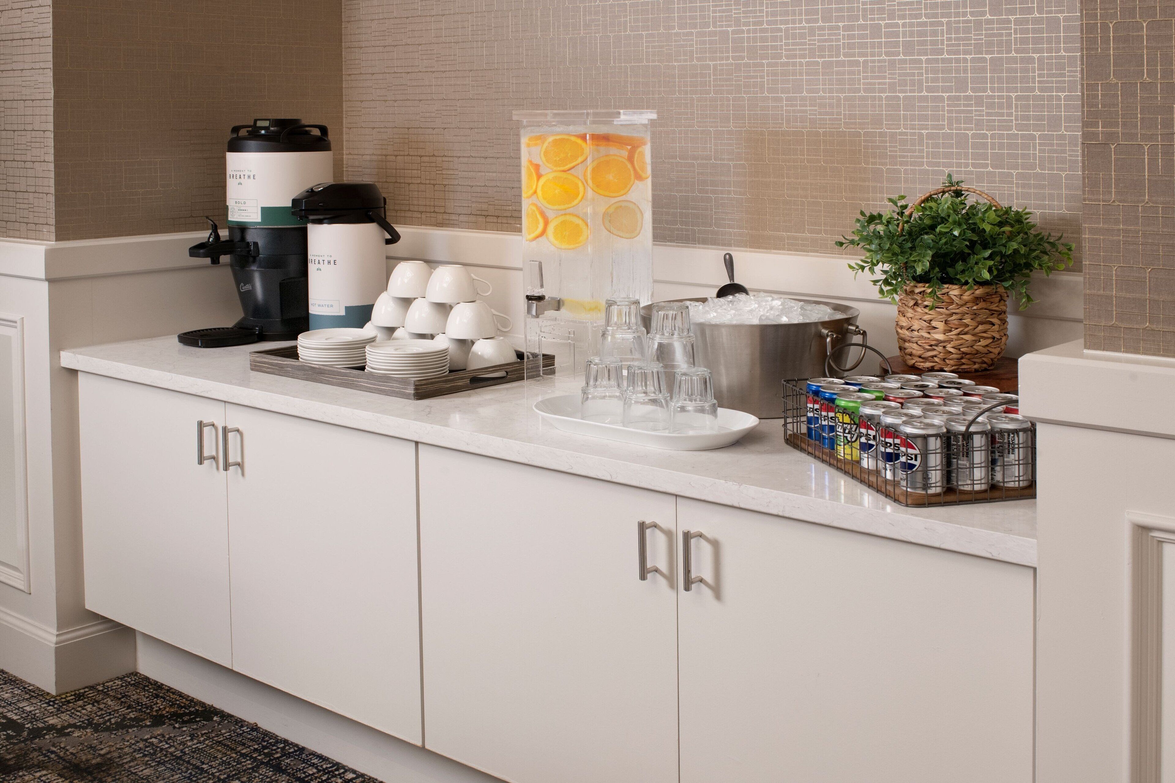 A hotel kitchen counter with a coffee maker, tea kettle, fruit-infused water dispenser, and a basket of fresh produce.