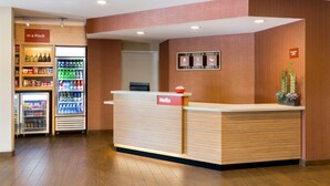 A hotel reception area with a wooden reception desk, a 'Hello' sign, a 'Stay' sign, a 'Work' sign, and a 'Play' sign. There are two refrigerators stocked with beverages and snacks.