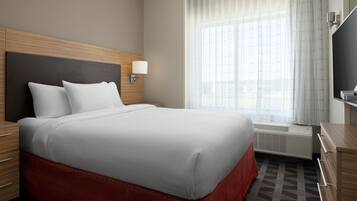 Habitación de hotel con una cama grande, cabecero de madera, mesita de noche con lámpara, ventana amplia con cortinas translúcidas y televisor de pantalla plana sobre un mueble de madera.