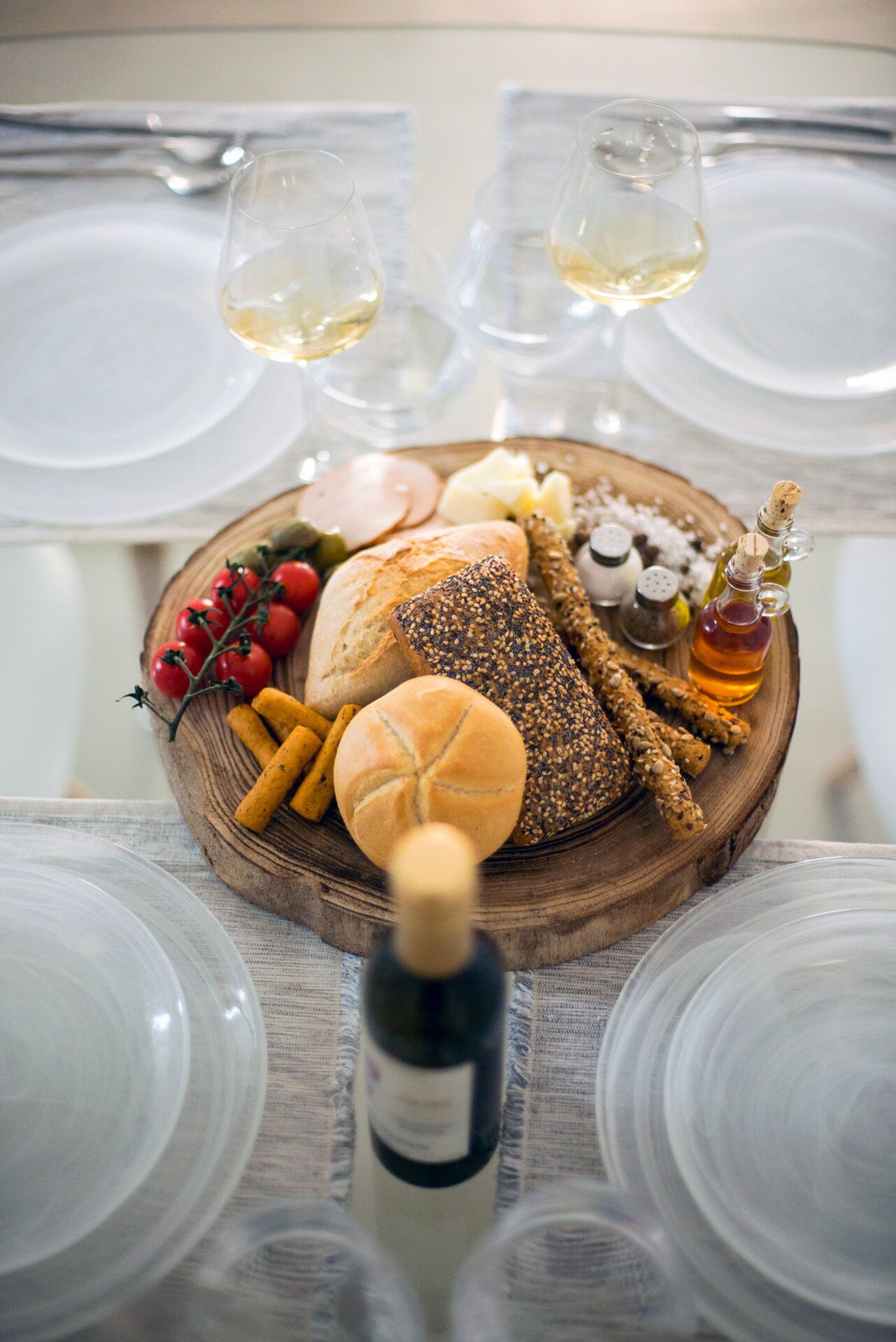 Una mesa dispuesta con una tabla de madera que sostiene varios panes, una botella de vino y condimentos.