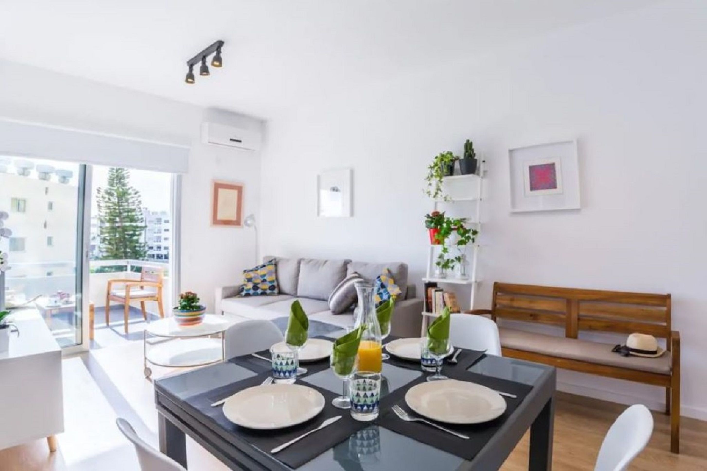 Un salon moderne avec un coin repas. La pièce est dotée d’une grande fenêtre, d’un canapé, d’une table basse et d’une table à manger, cette dernière étant mise avec des assiettes, des verres et une bouteille. On aperçoit des plantes sur une étagère et un banc sur lequel est posé un chapeau.