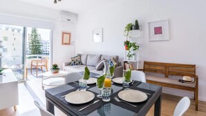 A modern living room with a dining area. The room has a large window, a sofa, a coffee table, and a dining table set with plates, glasses, and a bottle. There are plants on a shelf and a bench with a hat on it. - Marina's Flat Limassol (Limassol)